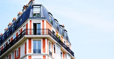 ein Apartment Hochhaus in rot und weiss gestrichen mit einem dunkelgrauem Dach, das vorletzte Stockwerk hat einen Balkon um das ganze Gebäude