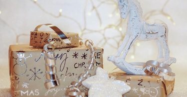 drei Geschenke in braunem Geschenkpapier mit silberner Schrift, Schneeflocken und Schneemännern, mit einem silbernem Band, einem kleinem weissen Schaukelpferd und im Hintergrund eine Lichterkette