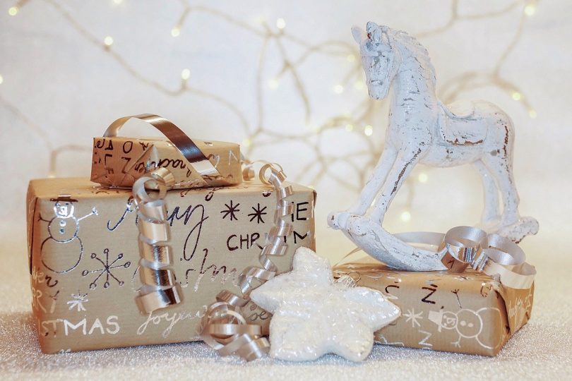 drei Geschenke in braunem Geschenkpapier mit silberner Schrift, Schneeflocken und Schneemännern, mit einem silbernem Band, einem kleinem weissen Schaukelpferd und im Hintergrund eine Lichterkette