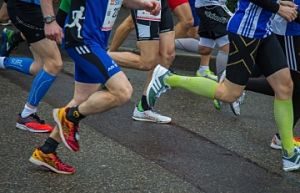 mehrere Renner auf einer Straße, einer mit blauem T-shirt und kurzer Hose,, und orangenen Turnschuhen und daneben einer in schwarzer kurzer Hose mit grünen Turnschuhen und grünen Kniestrümpfen