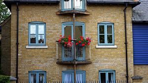 Ausschnitt eines Backsteinhauses mit blauen Fensterrahmen und in der Mitte ist ein jedem Stockwerk ein mini Balkon mit roten Geranien am Geländer