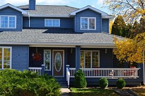 ein blaues zweistöckiges Einfamilienhaus mit niedrigen Hecken und grüner Wiese davor, am Eingang eine kleine Terrasse mit weissem Geländer