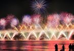 Feuerwerk am Hafen, unten sieht man helle Feuerwerksfontainen, darüber fünf runde Feuerwerke in lila und ganz oben wie ein lila Stern, im Vordergrund die Schatten vereinzelter Personen