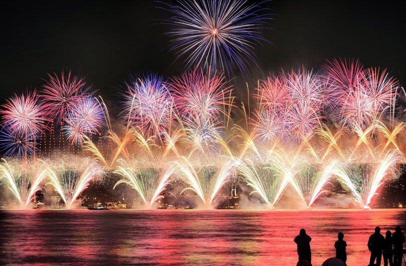Feuerwerk am Hafen, unten sieht man helle Feuerwerksfontainen, darüber fünf runde Feuerwerke in lila und ganz oben wie ein lila Stern, im Vordergrund die Schatten vereinzelter Personen