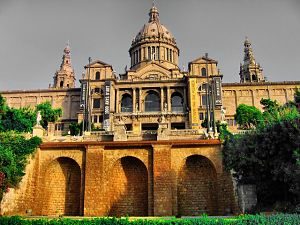 das nationale Kunstmuseum von Barcelona, man sieht von vorne das ganze Gebäude mit der Kuppel in der Mitte und zwei türmen links und rechts, davor grüne Wiese und einige Blätterbäume