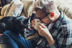 ein junger Mann mit schwarzer Brille, kariertem Hemd und Jeans der auf einem Sofa sitzt und aus einer Tasse Kaffee trinkt, eine graue Katze nähert sich ihm