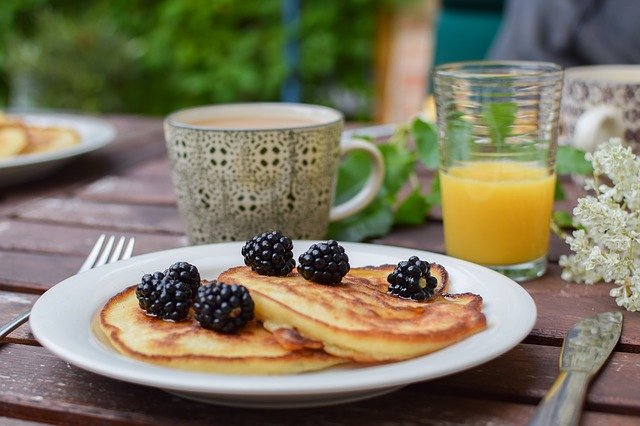 ein Holztisch mit einem weißen runden Teller auf dem zwei Pancakes mit Brombeeren liegen, daneben ein Glas halb voll mit Orangensaft und eine grüne Tasse