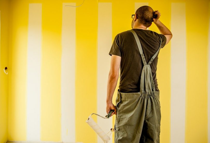 ein Mann in grauer Latzhose der ein der linken Hand einen Wandroller hält und sich mit der rechten Hand am Kopf kratzt, er steht vor einer gelb weiss gestreiften Wand in einer Wohnung