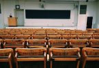 ein leeres Klassenzimmer einer Universität, ganz vorne eine lange Tafel und ein Lehrerpult und der erst des Raumes ist mit klappbaren Holzstühlen und Tischen gefüllt