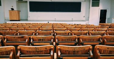 ein leeres Klassenzimmer einer Universität, ganz vorne eine lange Tafel und ein Lehrerpult und der erst des Raumes ist mit klappbaren Holzstühlen und Tischen gefüllt