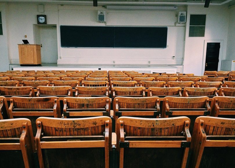 ein leeres Klassenzimmer einer Universität, ganz vorne eine lange Tafel und ein Lehrerpult und der erst des Raumes ist mit klappbaren Holzstühlen und Tischen gefüllt