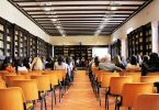Versammlung in einem länglichen Sal mit Bücherregalen an allen Wänden, bis auf einen Gang in der Mitte steht alles voller orangener Stühle, von denen nur die Hälfte von Personen besetzt sind und am Ende der Raumes ein Tisch en dem drei Personen sitzen