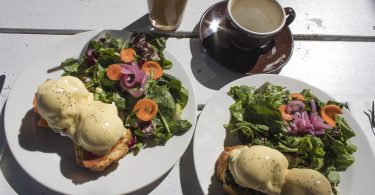 auf einem Holztisch stehen ein Latte Macchiato, ein Milchkaffee und zwei Teller mit zwei Benedict Eier auf Brot und daneben eingroßer Salat