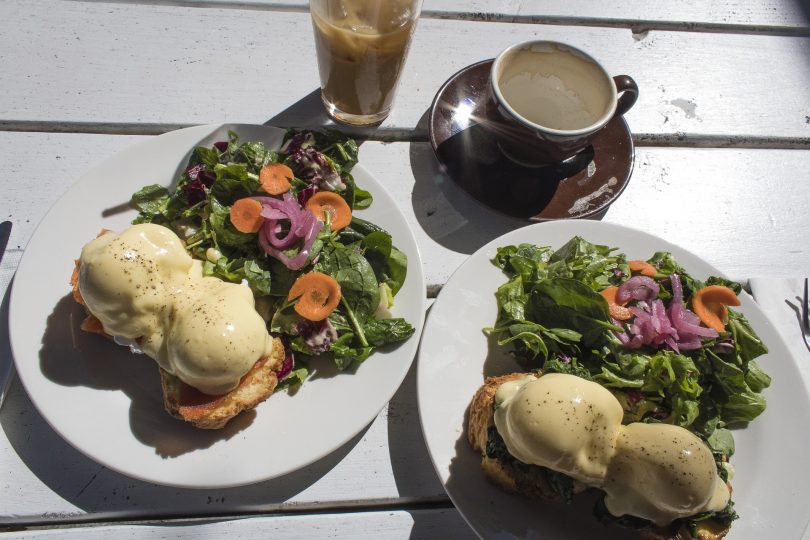 auf einem Holztisch stehen ein Latte Macchiato, ein Milchkaffee und zwei Teller mit zwei Benedict Eier auf Brot und daneben eingroßer Salat