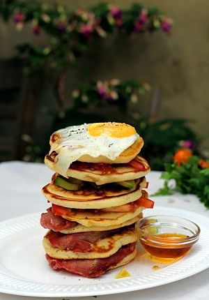 ein großer weisser Teller mit einem Pancaketurm oben drauf ein Spiegelei und zwischen den Törtchen Schinken und Tomate und daneben ein kleines Glasschüsselchen mit Ahornsirup