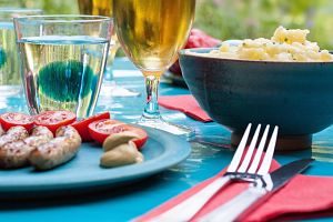 auf einem Tisch mit blauer Tischdecke zwei gefüllte Biergläser, eine Schüssel mit Kartoffelsalat, ein Glas Wasser, auf einer roten Serviette eine Gabel und ein Messer und ein Teller mit Würstchen und Tomatenscheiben