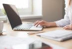 hölzerner Schreibtisch mit einem Laptop, einem Bleistift, mehrere Unterlagen und eine weibliche Hand die etwas am computer macht