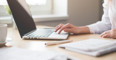 hölzerner Schreibtisch mit einem Laptop, einem Bleistift, mehrere Unterlagen und eine weibliche Hand die etwas am computer macht