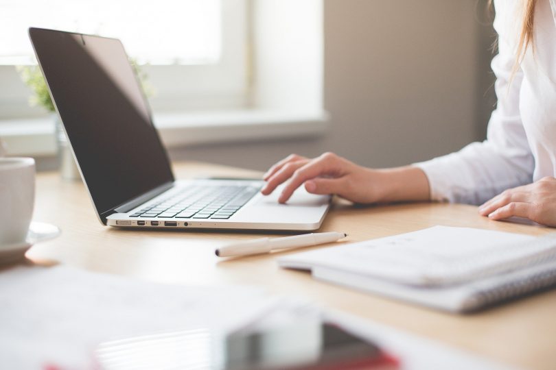 hölzerner Schreibtisch mit einem Laptop, einem Bleistift, mehrere Unterlagen und eine weibliche Hand die etwas am computer macht