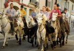 vier Männer und zwei Frauen die auf weißen und braunen Pferden durch die Straße reiten