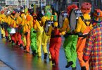 ein Faschingsumzug die Clowns laufen in einer Reihe mit ihren Musikinstrumenten alle mit orangnen Pullovern und grünen, roten oder gelben Hosen