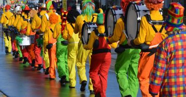 ein Faschingsumzug die Clowns laufen in einer Reihe mit ihren Musikinstrumenten alle mit orangnen Pullovern und grünen, roten oder gelben Hosen