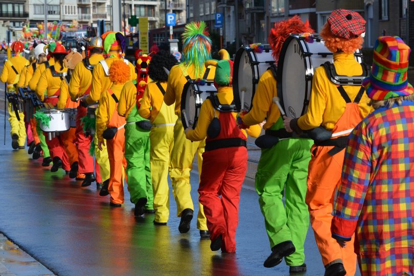 ein Faschingsumzug die Clowns laufen in einer Reihe mit ihren Musikinstrumenten alle mit orangnen Pullovern und grünen, roten oder gelben Hosen