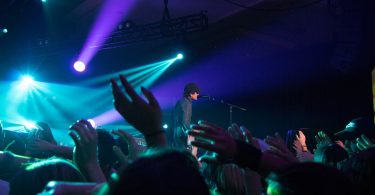 im Vordergrund eine Menschenmenge und auf der Bühne ein schwarzbekleideter Sänger mit Mütze türquises und Lianes Scheinwerferlicht