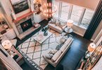 ein Wohnzimmer von oben fotografiert mit einem großen Teppich in der Mieter darauf ein beiges Sofa ein kleiner weißer viereckiger Tisch dahinter ein verschlossener Kamin und rechts Fenster über die ganze Wandbreite