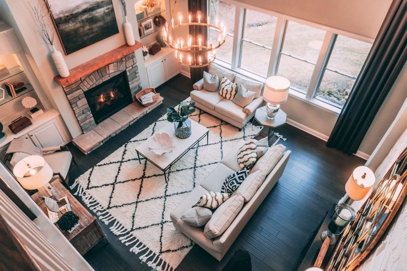 ein Wohnzimmer von oben fotografiert mit einem großen Teppich in der Mieter darauf ein beiges Sofa ein kleiner weißer viereckiger Tisch dahinter ein verschlossener Kamin und rechts Fenster über die ganze Wandbreite