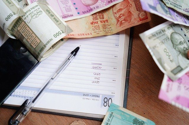 ein offenes Buch mit einem Kugelschreiber darauf in dem mehrere zahlen untereinander notiert wurden und drum herum liegen verschiedene indische Geldscheine