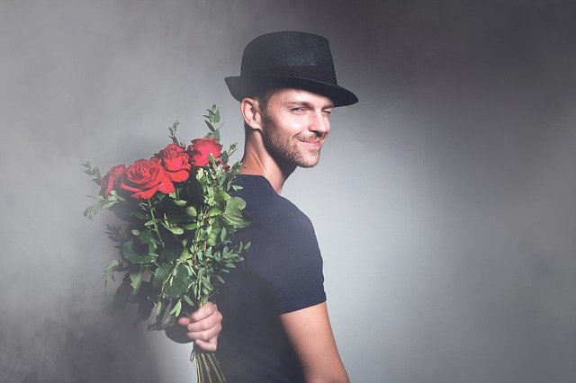 ein junger Mann im schwarzen T-Shirt, mit schwarzem Hut auf, der seitlich steht und lächelnd und Augenzwinkern nach hinten schaut und mit der linken Hand einen Strauß roter Rosen hinterm Rücken hält