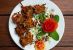 ein weißer runder Teller mit indischem Pakora auf der linken Seite und Salat mit minzblättern und einer in Rosenform geschnittene tomate