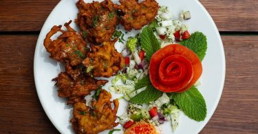 ein weißer runder Teller mit indischem Pakora auf der linken Seite und Salat mit minzblättern und einer in Rosenform geschnittene tomate