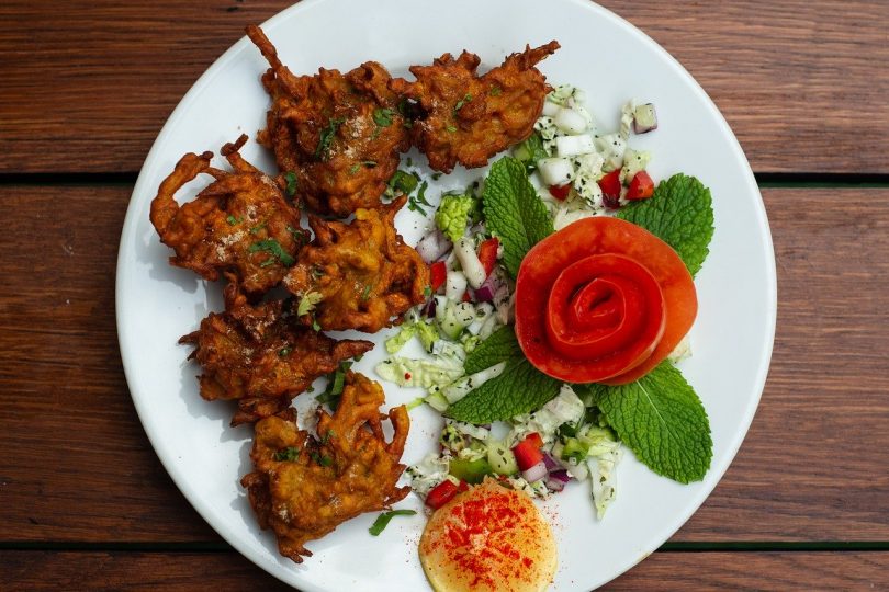 ein weißer runder Teller mit indischem Pakora auf der linken Seite und Salat mit minzblättern und einer in Rosenform geschnittene tomate