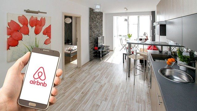 ein Wohnesszimmer im Hintergrund und vorne links im Bild eine Hand mit einem Handy auf dem die Seite Airbnb zu sehen ist