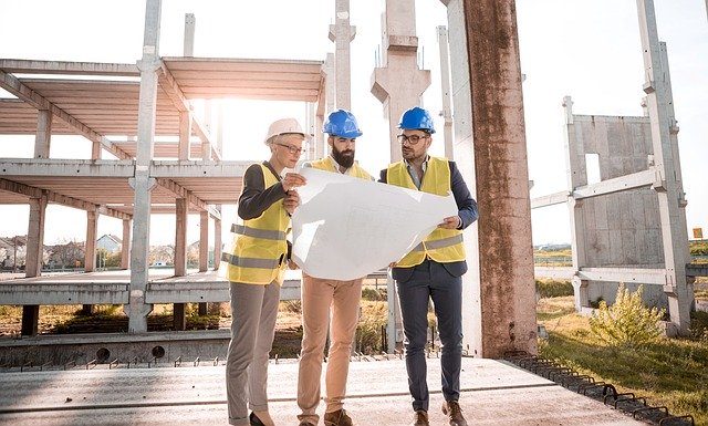 drei Architekten die auf einer Baustelle auf einen plan sehen, alle haben einen Helm auf und eine gelbe Weste an