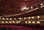 ein leeres Theater von innen, zu sehen sind die samtroten sitze und die verzierte Decke mit Lichtern
