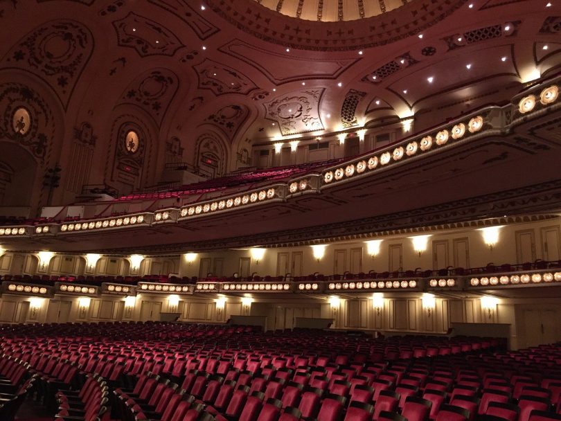 ein leeres Theater von innen, zu sehen sind die samtroten sitze und die verzierte Decke mit Lichtern