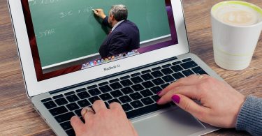 eine Person die an einem Laptop etwas schreibt während sie ein video von einem Lehrer sieht der eine mathematische Operation erklärt