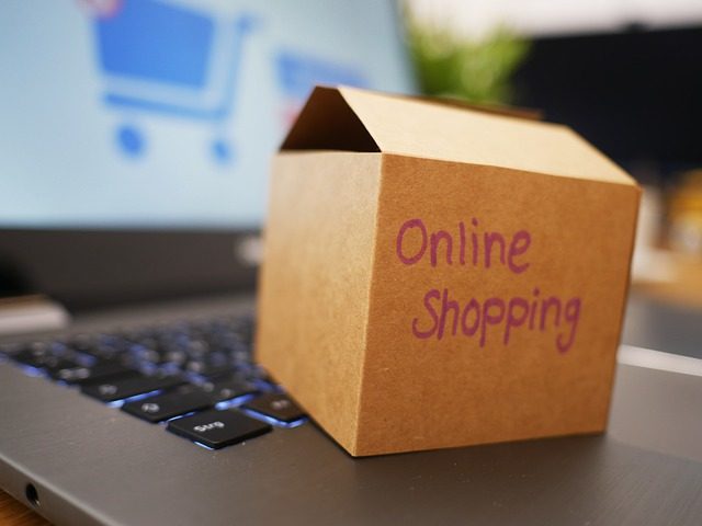 ein offener eingeschalteter Laptop mit einer kleinen Kartonkiste auf der Tastatur auf der mit lilanem Stift online shopping geschrieben steht