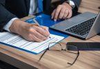 die arme eines Mannes im Anzug mit weißem Hemd und blauer Krawatte wie er gerade einen vertrag ausfüllt, vor ihm liegen eine Brille, ein Handy und ein Laptop auf einem Holztisch
