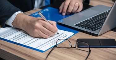 die arme eines Mannes im Anzug mit weißem Hemd und blauer Krawatte wie er gerade einen vertrag ausfüllt, vor ihm liegen eine Brille, ein Handy und ein Laptop auf einem Holztisch