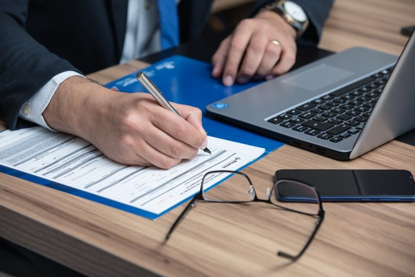 die arme eines Mannes im Anzug mit weißem Hemd und blauer Krawatte wie er gerade einen vertrag ausfüllt, vor ihm liegen eine Brille, ein Handy und ein Laptop auf einem Holztisch