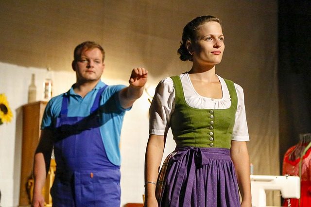 zwei Schauspieler die auf der Theaterbühne ein Eheleben darstellen, vorne eine Frau im Kleid und dahinter ein mann im blauen Arbeitsanzug der den Arm nach ihr ausstreckt