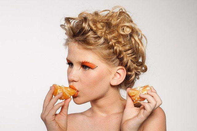 eine Frau mit orange geschminkten Augen, einen nach oben geflüchteten Zopf in ihren dunkelblonden Haar, die eine Mandarine isst