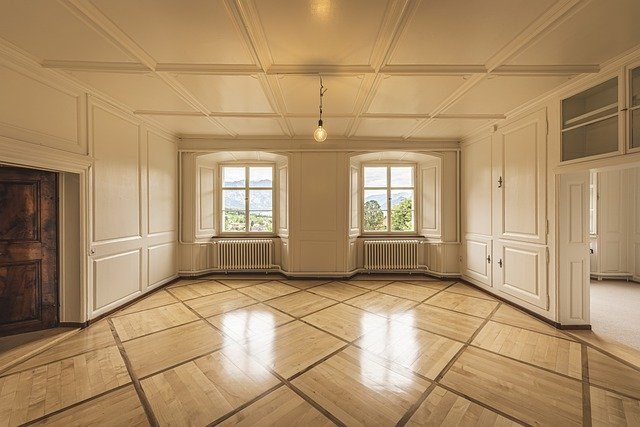 das Wohnzimmer einer unmöblierten Wohnung mit zwei großen Fenstern und links einem Kamin