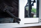 eine dunkel gekleidete Person die mit Hilfe eines Eisenstabs ein Fenster auf macht