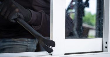 eine dunkel gekleidete Person die mit Hilfe eines Eisenstabs ein Fenster auf macht