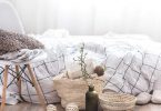 vor einem Bett mit karrierter Bettwäsche steht ein Holzhocker mit einem Kissen und einer Decke und am Boden zwei Weidenkörbe und eine grüne Flasche mit einem Zweig